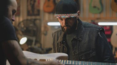 Şık bir atölyede elektro gitar yapan orta yaşlı marangozları kapatın. Profesyonel dövmeli bir zanaatkar iş arkadaşıyla konuşur ve gitar panosunu dener. El işi ve küçük işletme konsepti.