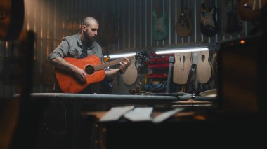 Erkek zanaatkar akustik gitar çalar, sesi kontrol eder. Ahşap ustası, marangoz modern atölyede ahşap müzik aleti yapıyor. Meslektaşlar arka planda çalışır. El yapımı ve girişimcilik..
