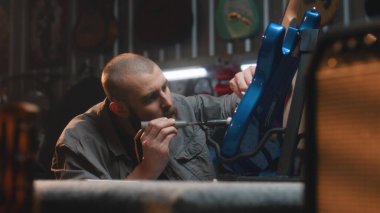 Profesyonel marangoz lehimleme demiri kullanarak gitar telleri çalar. Erkek zanaatkar modern atölyede ahşap müzik aleti yapıyor. İş arkadaşı arka planda çalışıyor. El yapımı ve girişimcilik..