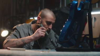 Profesyonel marangoz lehimleme demiri kullanarak gitar telleri çalar. Erkek zanaatkar modern atölyede ahşap müzik aleti yapıyor. İş arkadaşı arka planda çalışıyor. El yapımı ve girişimcilik..