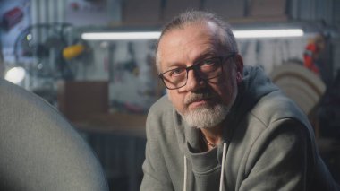 Gözlüklü profesyonel marangoz kameraya bakar. Erkek zanaatkar atölyede ya da stüdyoda mobilya montajı, yapımı ya da restorasyonu için çalışır. El işi ve küçük işletme konsepti. Portre.