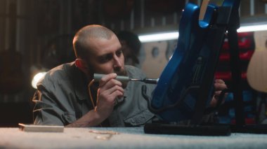 Profesyonel marangoz lehimleme demiri kullanarak gitar telleri çalar. Erkek zanaatkar modern atölyede ahşap müzik aleti yapıyor. İş arkadaşı arka planda çalışıyor. El yapımı ve girişimcilik..