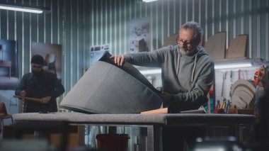 Olgun marangoz, mobilya yapımı ve restorasyonunda zımba tabancasıyla sandalye döşemesi yapıyor. Erkek meslektaşı arka planda rafları monte eder. El işi ve küçük işletmeler..