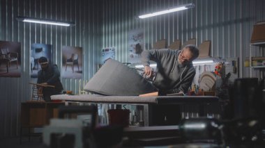 Olgun marangoz, mobilya yapımı ve restorasyonunda zımba tabancasıyla sandalye döşemesi yapıyor. Erkek meslektaşı arka planda rafları monte eder. El işi ve küçük işletmeler..