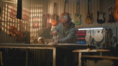 Olgun bir zanaatkar, ahşap gitar gövdesini cilalama makinesiyle parlatır. Ahşap ustası modern atölyede müzik aleti yapar. İş arkadaşı arka planda çalışıyor. El yapımı ve girişimcilik..