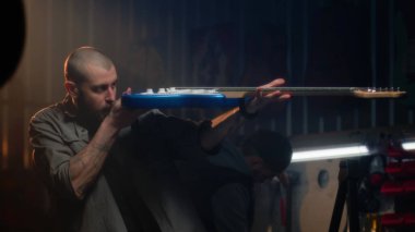 Erkek marangoz gitarı detaylıca inceler ve bez parçasıyla siler. Sanatçı modern atölyede ahşap müzik aleti yapıyor. İş arkadaşı arka planda çalışıyor. El işi ve küçük işletme konsepti.