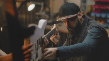 Şık bir atölyede elektro gitar yapan orta yaşlı marangozları kapatın. Profesyonel dövmeli bir zanaatkar iş arkadaşıyla konuşur ve gitar panosunu dener. El işi ve küçük işletme konsepti.