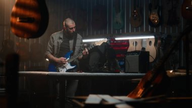 Erkek zanaatkar, kombinasyon amfisine bağlı gitar çalar, sesi kontrol eder. Marangoz modern atölyede ahşap müzik aleti yapıyor. Arka planda başka bir zanaatkar çalışıyor. El yapımı ve girişimcilik..