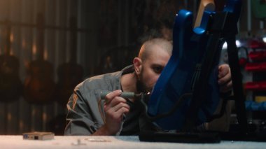 Profesyonel marangoz lehimleme demiri kullanarak gitar telleri çalar. Erkek zanaatkar modern atölyede ahşap müzik aleti yapıyor. İş arkadaşı arka planda çalışıyor. El yapımı ve girişimcilik..