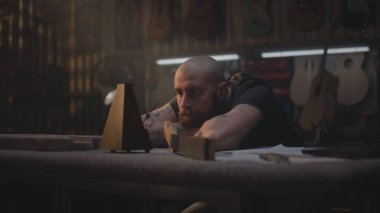 Yorgun marangoz metronoma bakar, müzik aletleri yapmak için atölyeye oturur. Erkek zanaatkar, zanaatkar uzun bir iş gününden sonra rahatlar. Duvarda tahta gitarlar asılı duruyor. El işi ve küçük işletmeler..