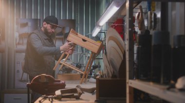 Erkek marangoz ahşap komodini yapar ya da monte eder. Genç bir zanaatkar, modern mobilya atölyesinde çalışıyor. Duvarında alet ve teçhizat olan tahta. El yapımı ve girişimcilik kavramı.