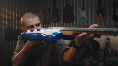 Erkek zanaatkar, bez parçasıyla ellerini siler, gitarı detaylıca inceler. Ahşap ustası modern atölyede ahşap müzik aleti yapıyor. İş arkadaşı arka planda çalışıyor. El yapımı ve girişimcilik..