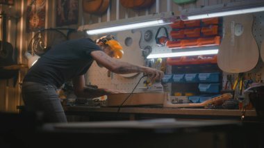 Koruyucu yüz kalkanı olan erkek marangoz, parlatma makinesi kullanarak tahta gitar gövdesini parlatır. Ahşap ustası, zanaatkar modern atölyede müzik aleti yapıyor. El yapımı ve girişimcilik..