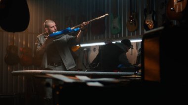 Erkek marangoz gitar tellerini bez parçasıyla siliyor ve gitarı inceliyor. Ahşap ustası, tasarımcı modern atölyede ahşaptan müzik aleti yapıyor. El işi ve küçük işletme konsepti.