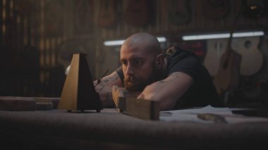 Yorgun marangoz metronoma bakar, müzik aletleri yapmak için atölyeye oturur. Erkek zanaatkar, zanaatkar uzun bir iş gününden sonra rahatlar. Duvarda tahta gitarlar asılı duruyor. El işi ve küçük işletmeler..