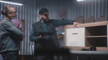 Profesyonel erkek tasarımcı dijital tablet bilgisayar kullanır, olgun marangoz veya zanaatkarla konuşur, ahşap TV standı yapmayı tartışır. Modern mobilya atölyesi. El yapımı ve girişimcilik kavramı.