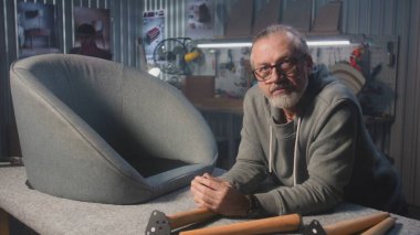 Profesyonel erkek marangoz sandalyeye yakın duruyor ve kameraya bakıyor. Olgun bir zanaatkar atölyede ya da stüdyoda montaj, üretim ya da yenileme mobilyaları için çalışır. El yapımı ve girişimcilikti. Portre.