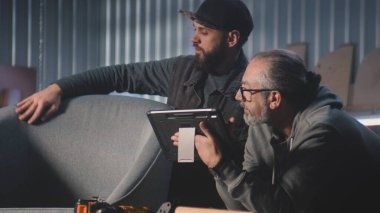 Olgun marangoz, tasarımcı iş arkadaşıyla konuşuyor, sandalye tasarımı ve yapımı hakkında konuşuyor, dijital tablet bilgisayar kullanıyor. Profesyonel zanaatkarlar şık mobilya atölyelerinde çalışırlar. El yapımı ve küçük işletmeler.