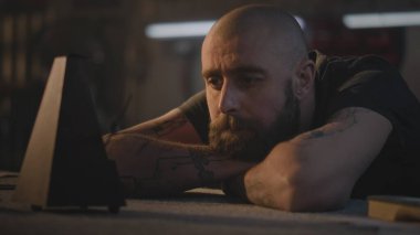 Orta yaşlı dövmeli bir zanaatkar işten sonra dinleniyor ve metronoma bakıyor. Profesyonel Kafkasyalı marangoz, karanlık atölyede masada oturur. El yapımı ve girişimcilik kavramı.