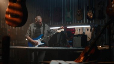 Erkek zanaatkar, kombinasyon amfisine bağlı gitar çalar, sesi kontrol eder. Marangoz modern atölyede ahşap müzik aleti yapıyor. Arka planda başka bir zanaatkar çalışıyor. El yapımı ve girişimcilik..