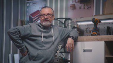 Gözlüklü olgun marangoz el yapımı ahşap TV standının yanında duruyor ve kameraya bakıyor. Profesyonel erkek zanaatkar. Mobilya yapmak için şık bir atölyede çalışıyor. Girişimcilik kavramı.