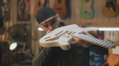 Şık bir atölyede elektro gitar yapan orta yaşlı marangozları kapatın. Profesyonel dövmeli bir zanaatkar iş arkadaşıyla konuşur ve gitar panosunu dener. El işi ve küçük işletme konsepti.