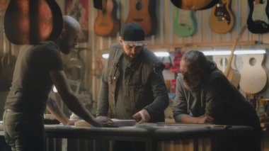 Olgun beyaz marangoz planlara bakar ve şık bir ışık atölyesinde çalışmayı tartışır. Erkek dövmeli zanaatkar gitar gövdesini tutuyor ve iş arkadaşlarıyla konuşuyor. El yapımı ve girişimcilik kavramı.