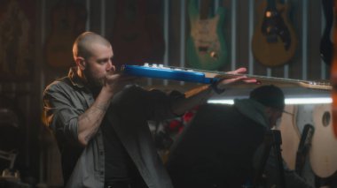 Erkek marangoz gitarı detaylıca inceler ve bez parçasıyla siler. Sanatçı modern atölyede ahşap müzik aleti yapıyor. İş arkadaşı arka planda çalışıyor. El işi ve küçük işletme konsepti.