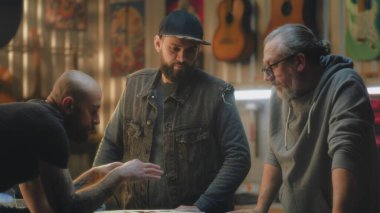 Erkek sanatçılar işi tartışıyor ve hafif stil atölyesinin planlarını kontrol ediyorlar. Dövmeli marangoz elektro gitar yapıyor ve iş arkadaşlarıyla konuşuyor. El işi ve küçük işletme kavramı.