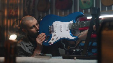 Erkek marangoz gitar ve telleri detaylı olarak inceler. Sanatçı modern atölyede ahşaptan müzik aleti yapıyor. Arka planda başka bir zanaatkar çalışıyor. El işi ve küçük işletme konsepti.