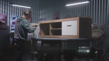 Olgun marangoz televizyon standını tornavida kullanarak monte eder. Arka planda başka bir uzman çalışıyor. İki zanaatkar mobilya yapmak için modern atölyede çalışıyor. El işi ve küçük işletme konsepti.