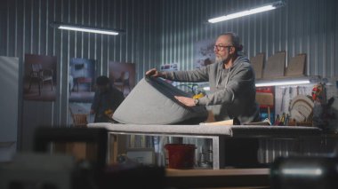 Olgun marangoz, mobilya yapımı ve restorasyonunda zımba tabancasıyla sandalye döşemesi yapıyor. Erkek meslektaşı arka planda rafları monte eder. El işi ve küçük işletmeler..