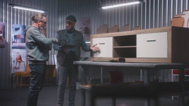 Profesyonel erkek marangoz ya da tasarımcı, dijital tablet bilgisayarı ile olgun iş arkadaşlarıyla konuşur ve ahşap TV standı yapmayı tartışır. Modern mobilya atölyesi. El işi ve küçük işletme konsepti.