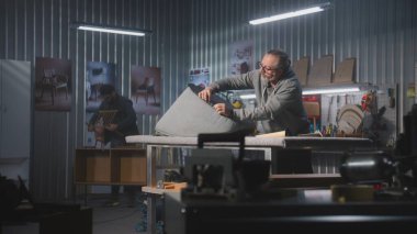 Olgun marangoz, mobilya yapımı ve restorasyonunda zımba tabancasıyla sandalye döşemesi yapıyor. Erkek meslektaşı arka planda rafları monte eder. El işi ve küçük işletmeler..