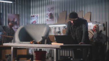 Erkek marangoz, tasarımcı dijital tablet ve dizüstü bilgisayar kullanıyor, modern atölyede mobilya yapımı ve yenilenmesi için sandalyeyle çalışıyor. Olgun uzman arka planda rafları birleştirir. El yapımı konsept.