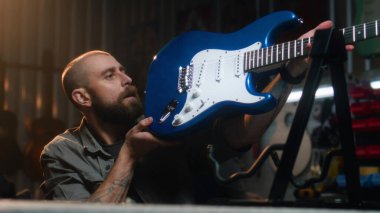 Erkek marangoz gitarı inceler ve ipleri paçavrayla siler. Ahşap işçiliği modern atölyede ahşap müzik aleti yapıyor. İş arkadaşı arka planda çalışıyor. El yapımı ve girişimcilik..