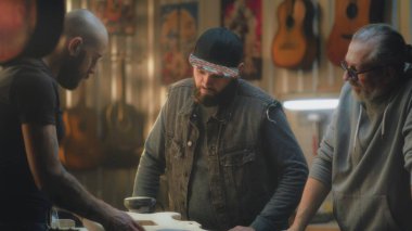 Erkek sanatçılar işi tartışıyor ve hafif stil atölyesinin planlarını kontrol ediyorlar. Dövmeli marangoz elektro gitar yapıyor ve iş arkadaşlarıyla konuşuyor. El işi ve küçük işletme kavramı.