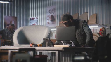 Erkek marangoz veya tasarımcı tablet bilgisayar ve dizüstü bilgisayar kullanarak hesaplamalar yapar. Zanaatkar, modern mobilya atölyesinde sandalye yapmak veya restore etmek için çalışıyor. El yapımı ve girişimcilik kavramı.