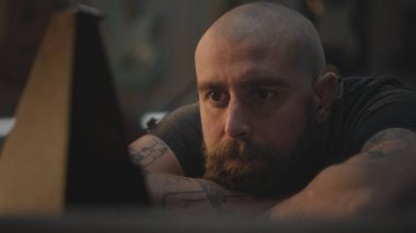 Orta yaşlı dövmeli bir zanaatkar işten sonra dinleniyor ve metronoma bakıyor. Profesyonel Kafkasyalı marangoz, karanlık atölyede masada oturur. El yapımı ve girişimcilik kavramı.