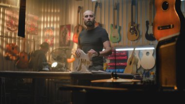 Erkek küçük işletme sahibi, girişimci modern marangoz atölyesinde duruyor, tahta gitar gövdesini elinde tutuyor ve kameraya bakıyor. Marangozlar ya da zanaatkarlar arka planda çalışır. El işi kavramı.