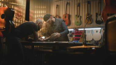 İki profesyonel marangoz, ahşap gitar gövdesini inceliyor ve modern atölyede müzik aleti yapma sürecini tartışıyorlar. İş arkadaşı arka planda çalışıyor. El yapımı ve girişimcilik kavramı.