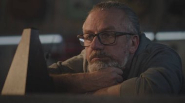 Olgun profesyonel bir zanaatkar karanlık bir atölyede masada oturur ve metronoma bakar. Yorgun marangoz dinlenir, gözlüğünü çıkarır ve ellerine uzanır. El yapımı ve girişimcilik kavramı.