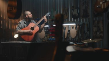 Erkek zanaatkar akustik gitar çalar, sesi kontrol eder. Ahşap ustası, marangoz modern atölyede ahşap müzik aleti yapıyor. Meslektaşlar arka planda çalışır. El yapımı ve girişimcilik..