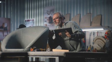 Olgun marangoz veya tasarımcı, şık bir atölyede mobilya yapımı ve restorasyonu için tablet bilgisayar kullanır. İş arkadaşı arka planda çalışıyor. El işi ve küçük işletmeler..