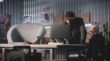 Erkek marangoz, tasarımcı dijital tablet ve dizüstü bilgisayar kullanıyor, modern atölyede mobilya yapımı ve yenilenmesi için sandalyeyle çalışıyor. Olgun uzman arka planda rafları birleştirir. El yapımı konsept.