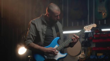 Erkek marangoz gitarı detaylıca inceler ve bez parçasıyla siler. Sanatçı modern atölyede ahşap müzik aleti yapıyor. İş arkadaşı arka planda çalışıyor. El işi ve küçük işletme konsepti.