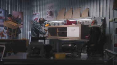 Olgun marangoz televizyon standını tornavida kullanarak monte eder. Başka bir erkek işçi komodin yapıyor. İki zanaatkar mobilya yapmak için modern atölyede çalışıyor. El yapımı ve girişimcilik kavramı.