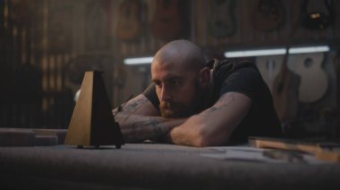 Yorgun marangoz metronoma bakar, müzik aletleri yapmak için atölyeye oturur. Erkek zanaatkar, zanaatkar uzun bir iş gününden sonra rahatlar. Duvarda tahta gitarlar asılı duruyor. El işi ve küçük işletmeler..