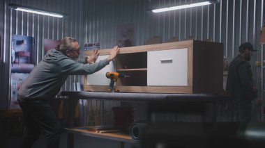 Olgun marangoz televizyon standını tornavida kullanarak monte eder. Arka planda başka bir uzman çalışıyor. İki zanaatkar mobilya yapmak için modern atölyede çalışıyor. El işi ve küçük işletme konsepti.