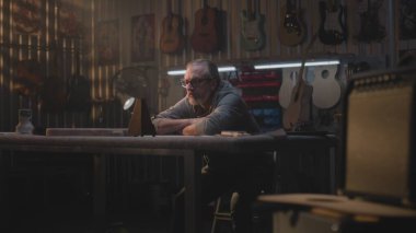 Yorgun erkek zanaatkar gözlüğünü çıkarıp metronoma bakıyor. Olgun marangoz müzik enstrümanları yapmak için modern atölyede uzun bir çalışma gününden sonra dinlenir. Duvarda el yapımı ahşap gitarlar asılı..